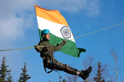 An Indian soldier in a blue helmet sliding along a rope while holding the tricolour flag against a blue sky