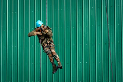A soldier in a blue helmet rappelling down a green metal wall