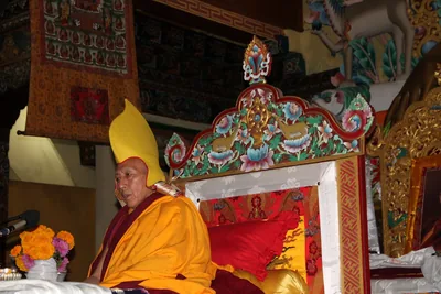 A senior Buddhist lama in a yellow hat seated beside a richly painted throne