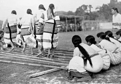 Old black and white photo of women in striped wraps stepping over long bamboo poles while others kneel