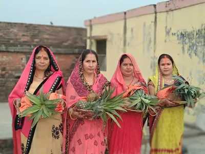 Women in bright saris holding pineapples and fruit offerings outside a house