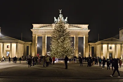 A tall lit Christmas tree in front of a grand stone gate at night, with people walking by