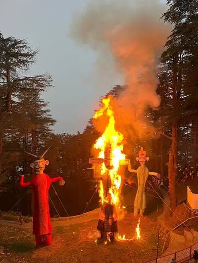 A tall effigy of Ravana burning in bright flames among pine trees, with two more effigies standing beside it