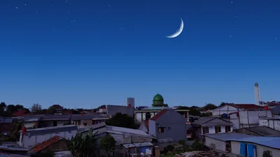 A thin crescent moon in a deep blue evening sky over rooftops and a green mosque dome