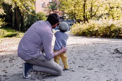 A father kneels on a park path beside his small son, seen from behind