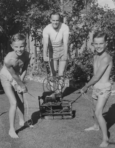 A black and white photo of a father pushing a lawnmower with his two sons in 1954