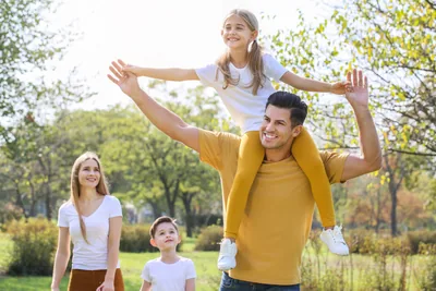 A father carrying his daughter on his shoulders in a sunny park while the family looks on