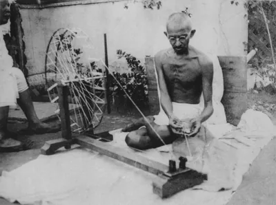 Black and white photo of Gandhi seated on the floor spinning thread at a wooden spinning wheel
