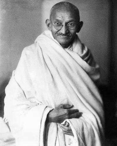 Black and white studio portrait of a smiling Gandhi wrapped in a white shawl
