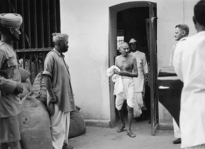 Black and white photo of Gandhi stepping out of a doorway while a man waits outside