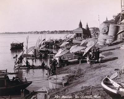 An old photograph of boats and umbrellas along a riverside ghat with temples