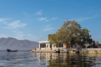 A small white temple at the edge of a lake under a big tree with hills behind