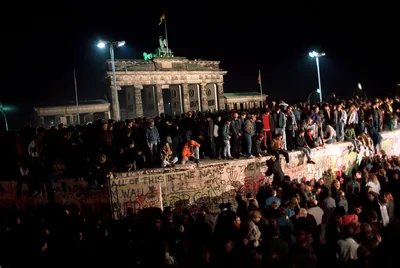 At night, a large crowd stands on and around the Berlin Wall in front of the lit Brandenburg Gate