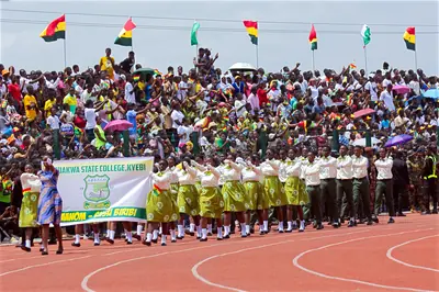 Rows of students in uniform march on a running track before stands full of people and flags