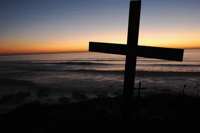 A wooden cross on a hilltop against an orange sunset over the sea