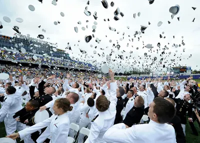 Hundreds of graduates in white uniforms throw their caps high into the air in a stadium