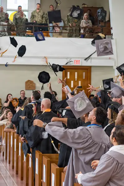 Graduates in gowns cheer and throw caps as families watch from the stands