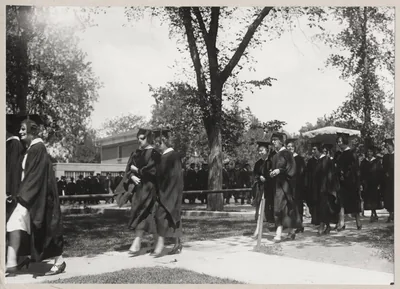 An old photo of graduates in gowns walking in a procession under tall trees
