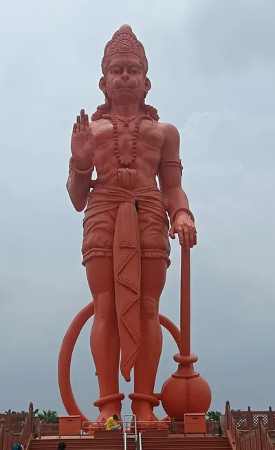 A tall red statue of Lord Hanuman raising his hand in blessing against the sky