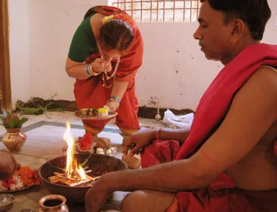 A priest and a woman in a red sari offering into a small sacred fire at a home ceremony