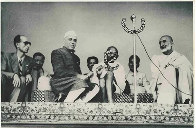 Black and white photo of Jawaharlal Nehru and Mahatma Gandhi seated on a stage before a microphone