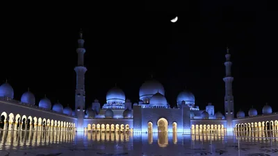 A white mosque with domes lit blue at night under a thin crescent moon