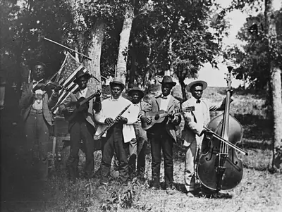 Old black and white photo of a band with brass instruments and a double bass under trees
