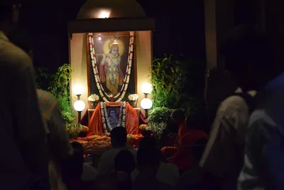 A shrine with a painting of Krishna, garlands and lamps, devotees sitting in the dark before it