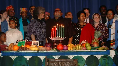 People in African print clothes stand behind a long table with a lit kinara, fruit and vegetables