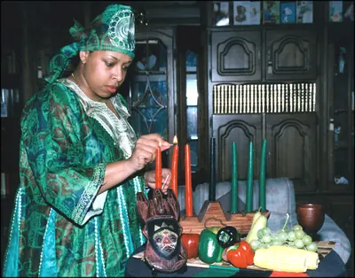 A woman in a green African print dress lights the candles of a kinara beside fruit and corn