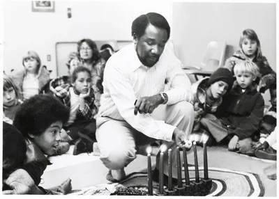 Black and white photo of a man kneeling to light a kinara as a group watches