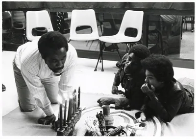 Black and white photo of a group seated on the floor around a kinara and a mat