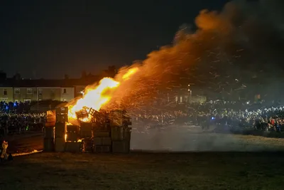 A large bonfire throwing sparks into the night sky with a crowd watching from a distance