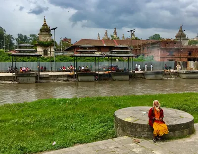 A sadhu in saffron robes sitting by a river with temples on the far bank
