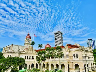 A grand building with a clock tower and copper domes under a blue sky, with the Malaysian flag