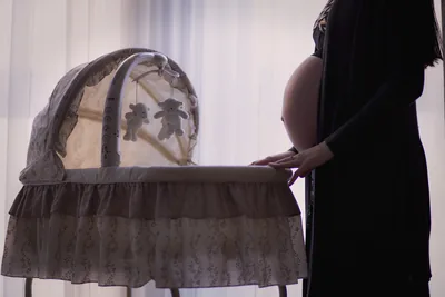 A mother-to-be stands beside a lace baby cradle by a window