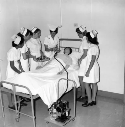 Trainee nurses in white caps gathered around a hospital bed