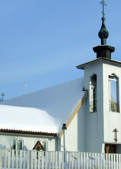 A white Orthodox church with a dark onion dome and a snowy roof under a blue sky