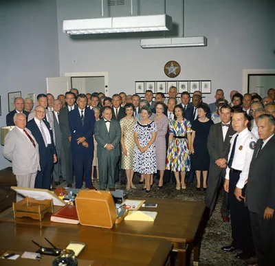 A large group of men and women in formal clothes gathered in an office for a retirement party in the 1960s