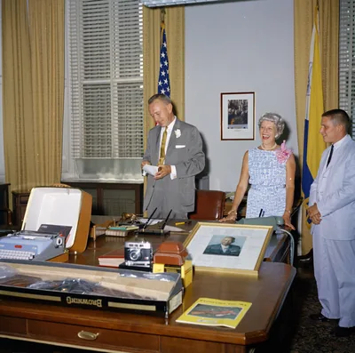 A retiring official stands by his desk with his wife and a colleague in an office