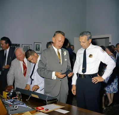 Men in suits and uniform gather around a desk at a farewell gathering