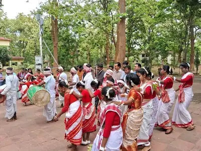 Women in red and white saris dance in a line under sal trees as drummers play