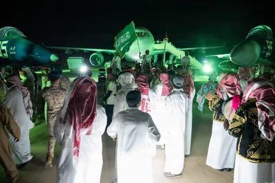 A crowd in white thobes waves green flags under green lights at night