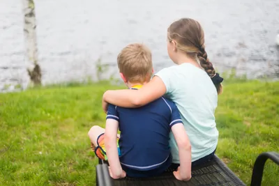 A boy and girl sitting on the grass by a lake, arm in arm