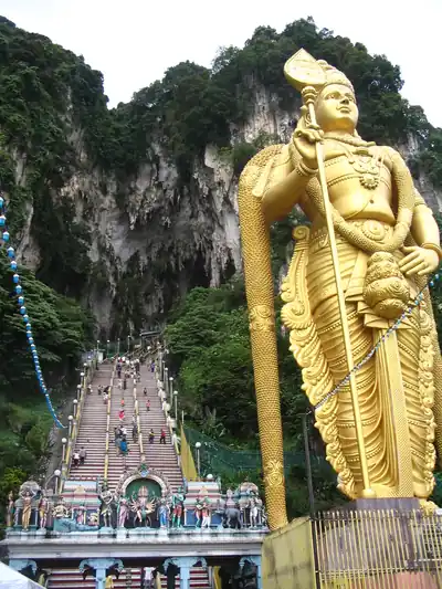 A giant golden statue of Murugan beside long stairs up to a limestone cave