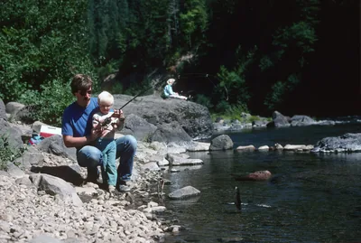 A father and son sitting on rocks fishing by a clear river
