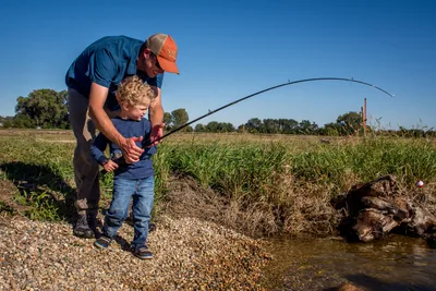 A father helping his young son hold a fishing rod on a river bank