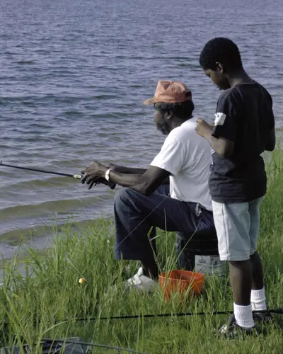 A father fishing from a grassy bank as his son stands beside him