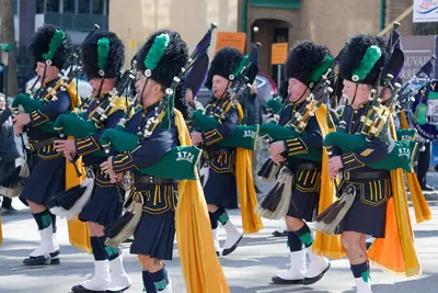 Pipers in tall black bearskin hats and green sashes marching in a parade