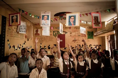 A classroom of children in uniform raising their hands eagerly with portraits on the wall